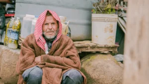 An elderly man sits on the ground, wearing a scarf, exuding a sense of wisdom and quiet dignity.