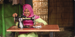 A woman wearing a pink headscarf skillfully sews fabric on a sewing machine, focused on her craft.