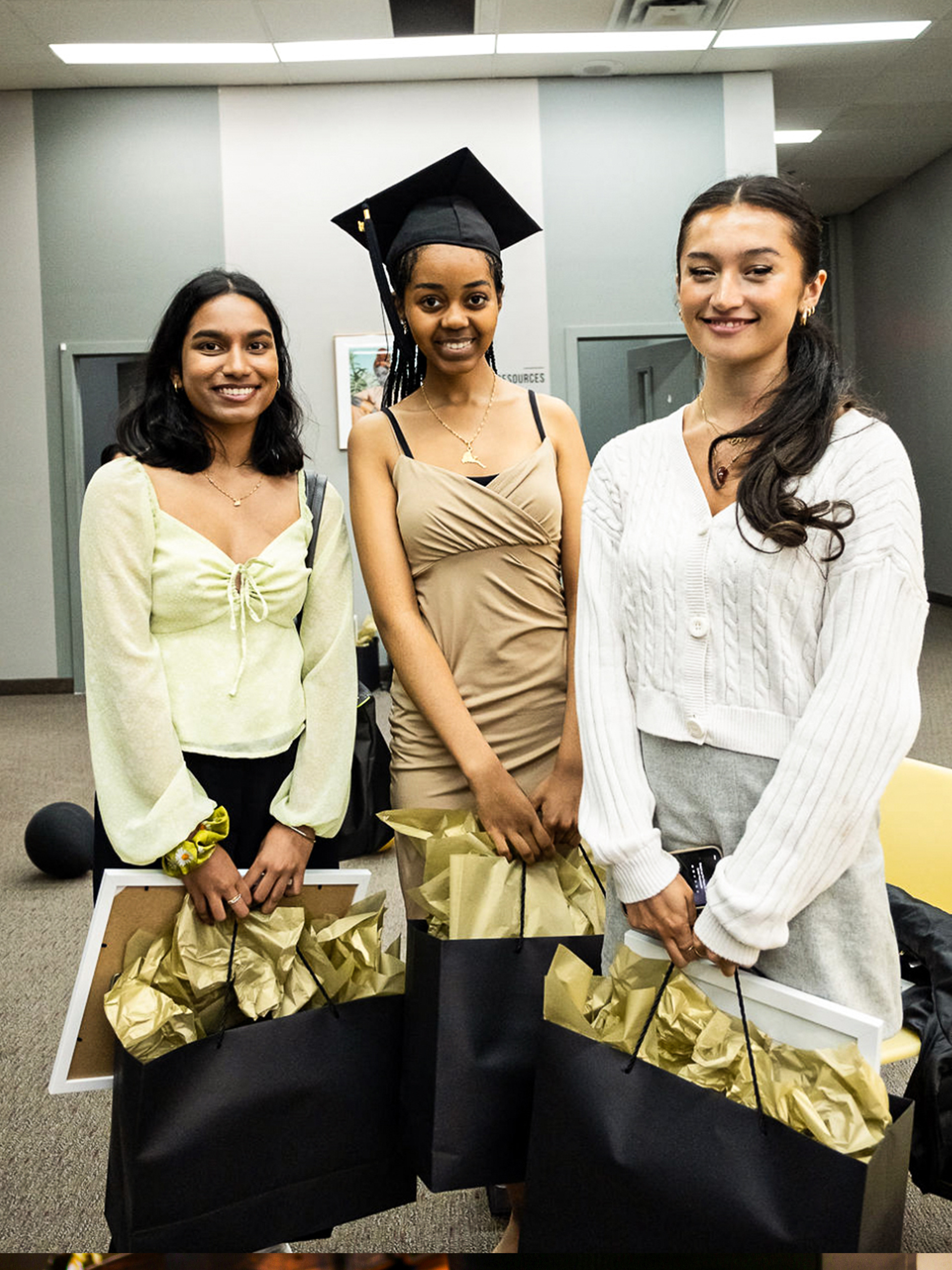 Three women in graduation gowns proudly hold their bags, celebrating their academic achievements together.