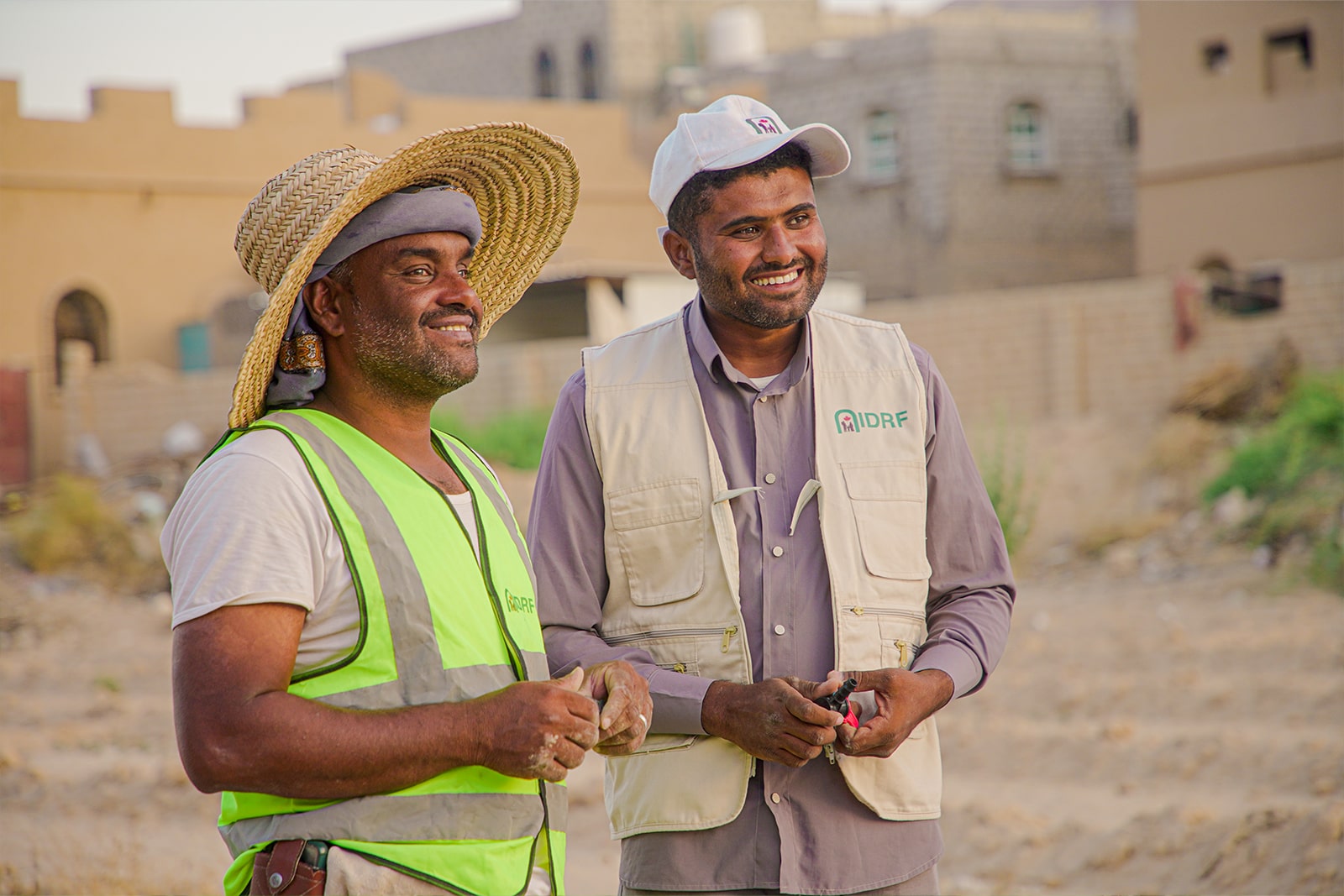 Two men in vests and hats stand on the sand, exuding a sense of camaraderie and dignity in their relaxed posture.
