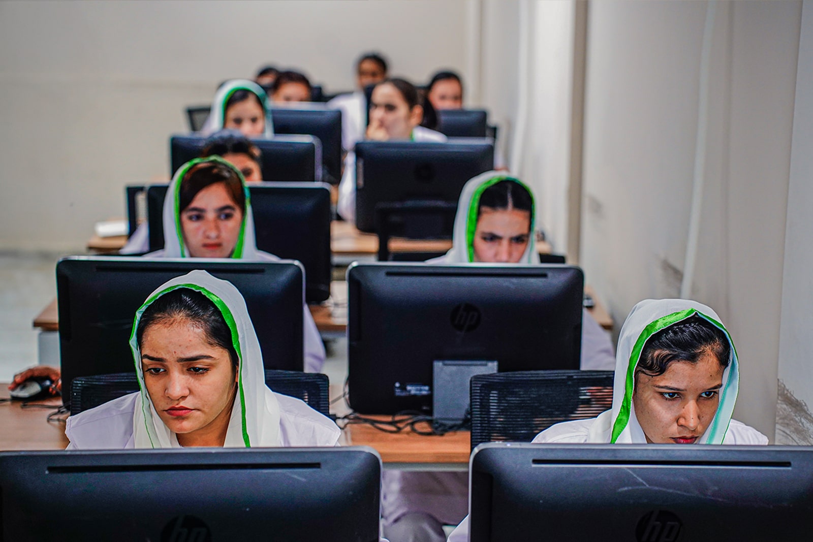 A group of women wearing white headscarves, focused on their work at computers in a collaborative environment.