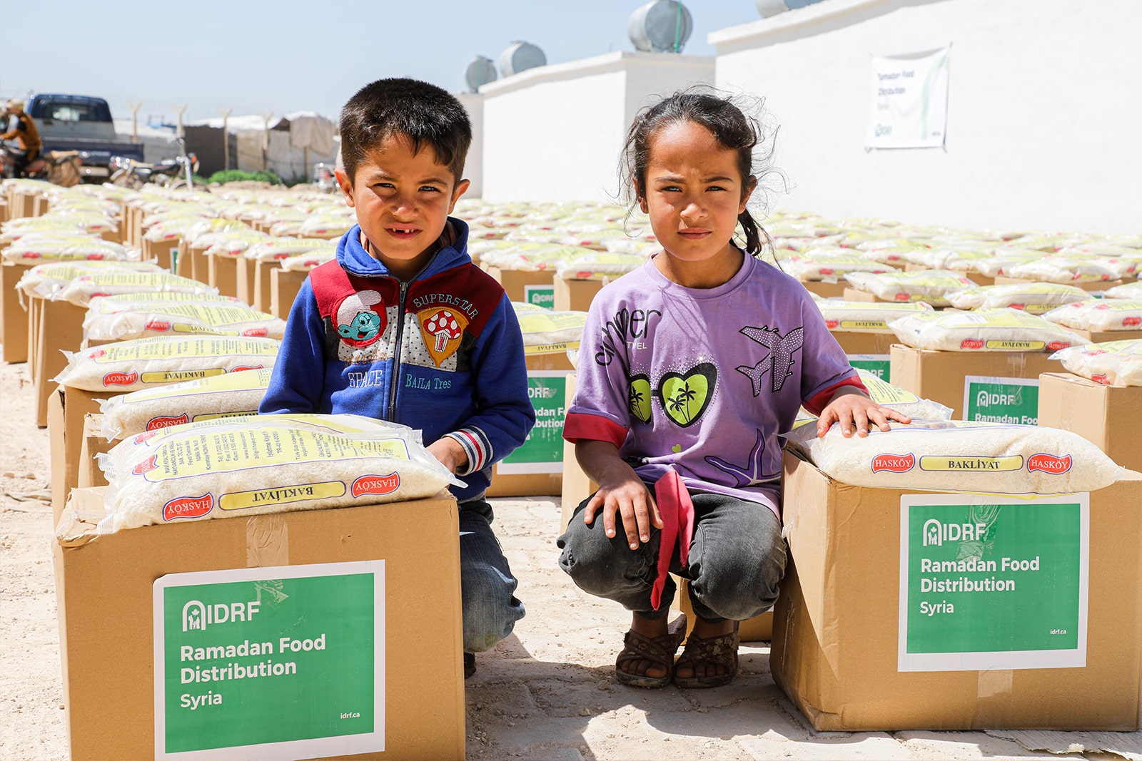 Two children sit on stacked food boxes, sharing a moment of camaraderie amidst a backdrop of community support.