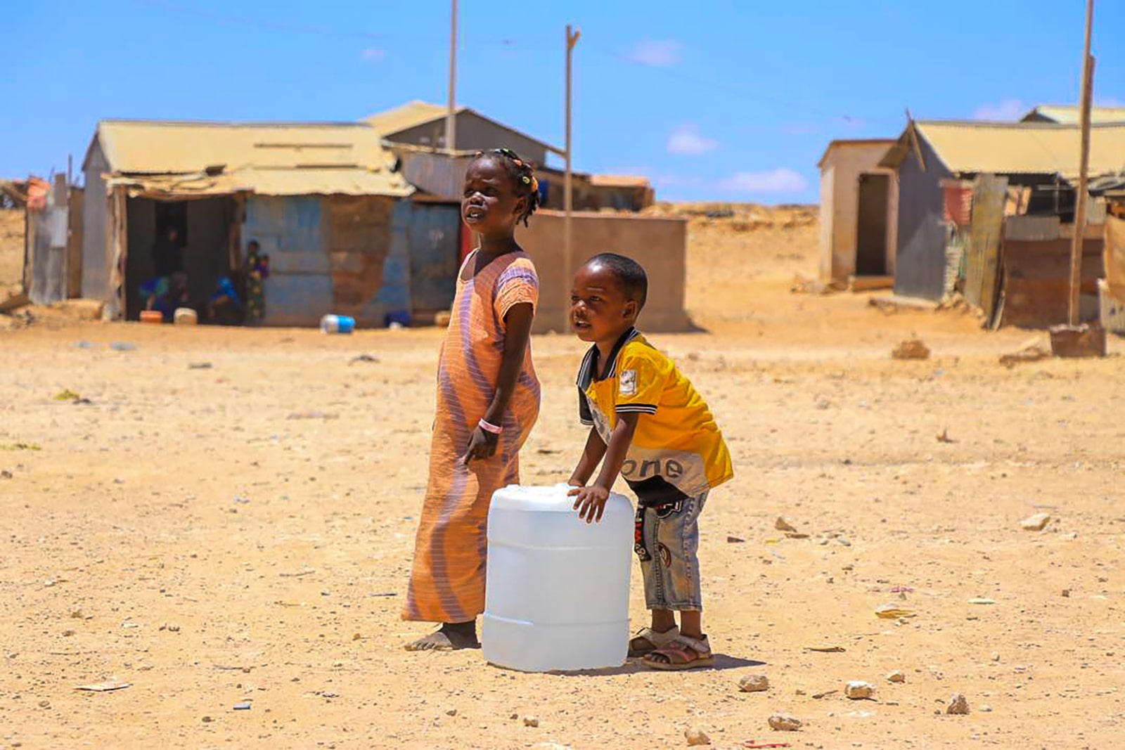 Two children stand beside a water container in a dusty field, highlighting the importance of water access in rural areas.