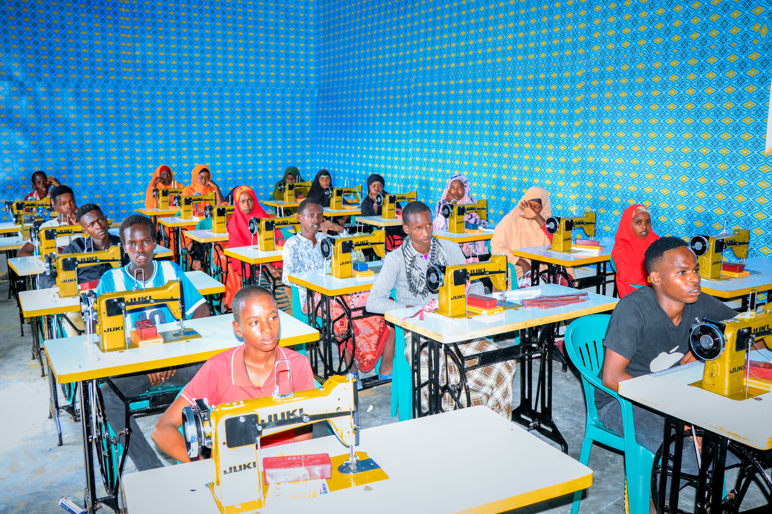 A group of teens focused on sewing at their desks, each using a sewing machine in a collaborative workspace.