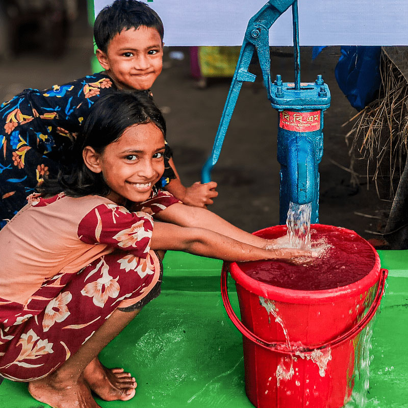 Two smiling children pump water into a red bucket from a blue hand pump. The scene is joyful and vibrant, with the girl washing her hands.