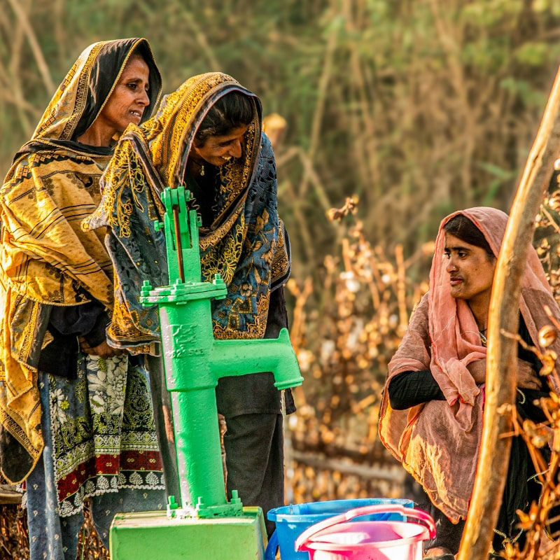 Three women wearing colorful shawls gather around a bright green hand pump. One woman tends the pump while the other two engage in conversation, with colorful buckets awaiting water. The setting is sunlit, suggesting an early morning or late afternoon scene, evoking a sense of community and daily routine.