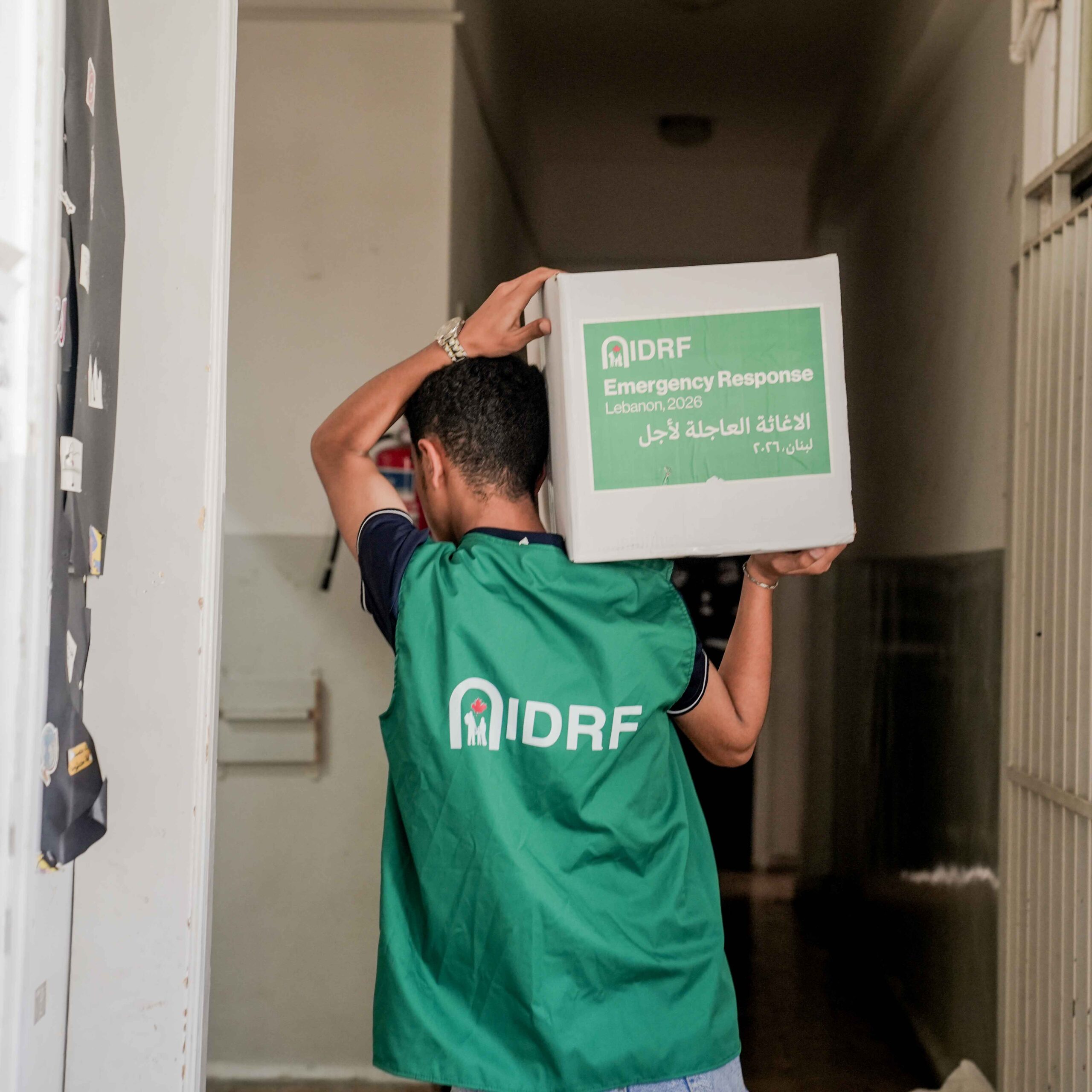 A person wearing a green IDRF vest carries a box labeled "Emergency Response Lebanon 2026" down a hallway.