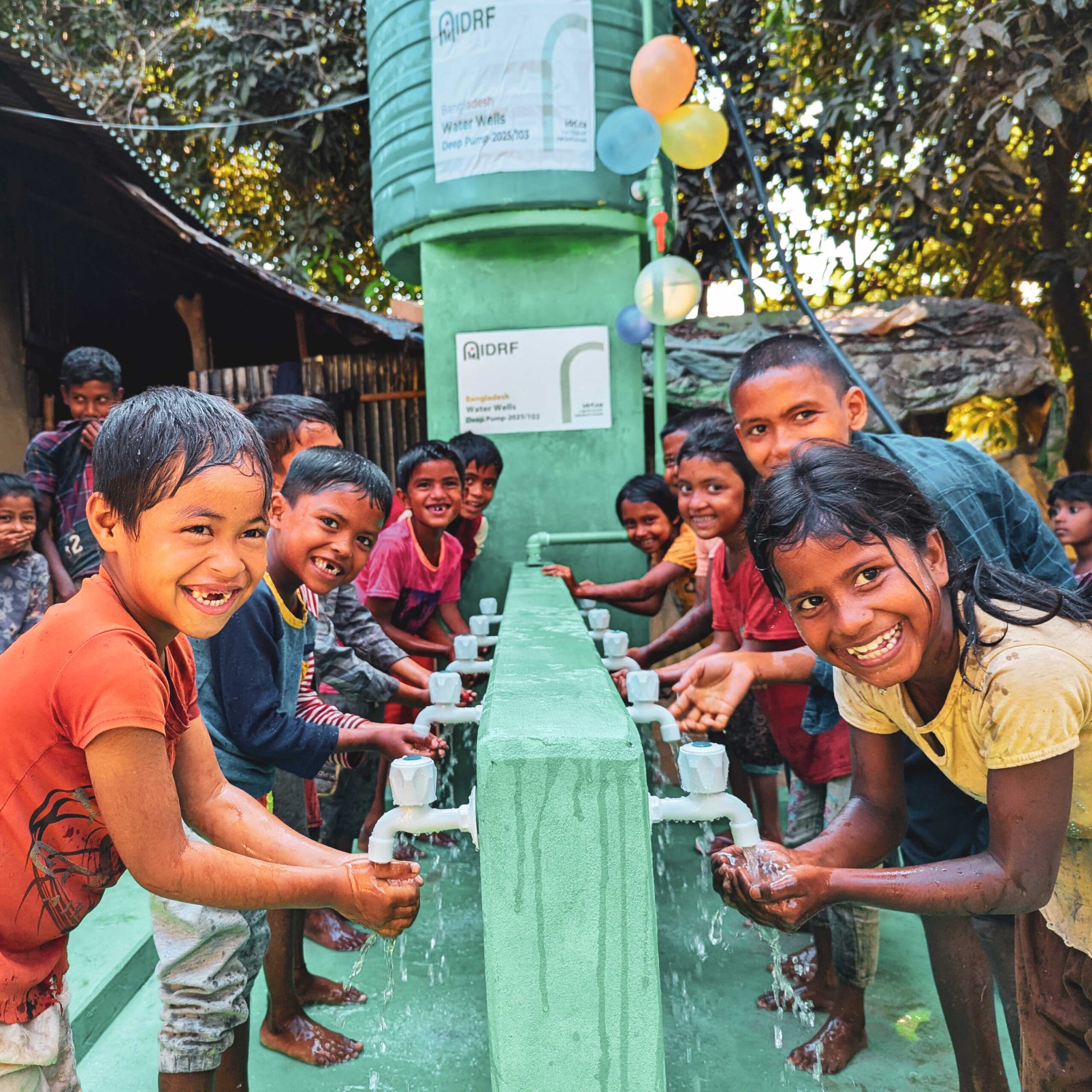 Smiling children gather around a water station, washing hands under flowing taps. The scene is vibrant and joyful, with balloons and greenery around.