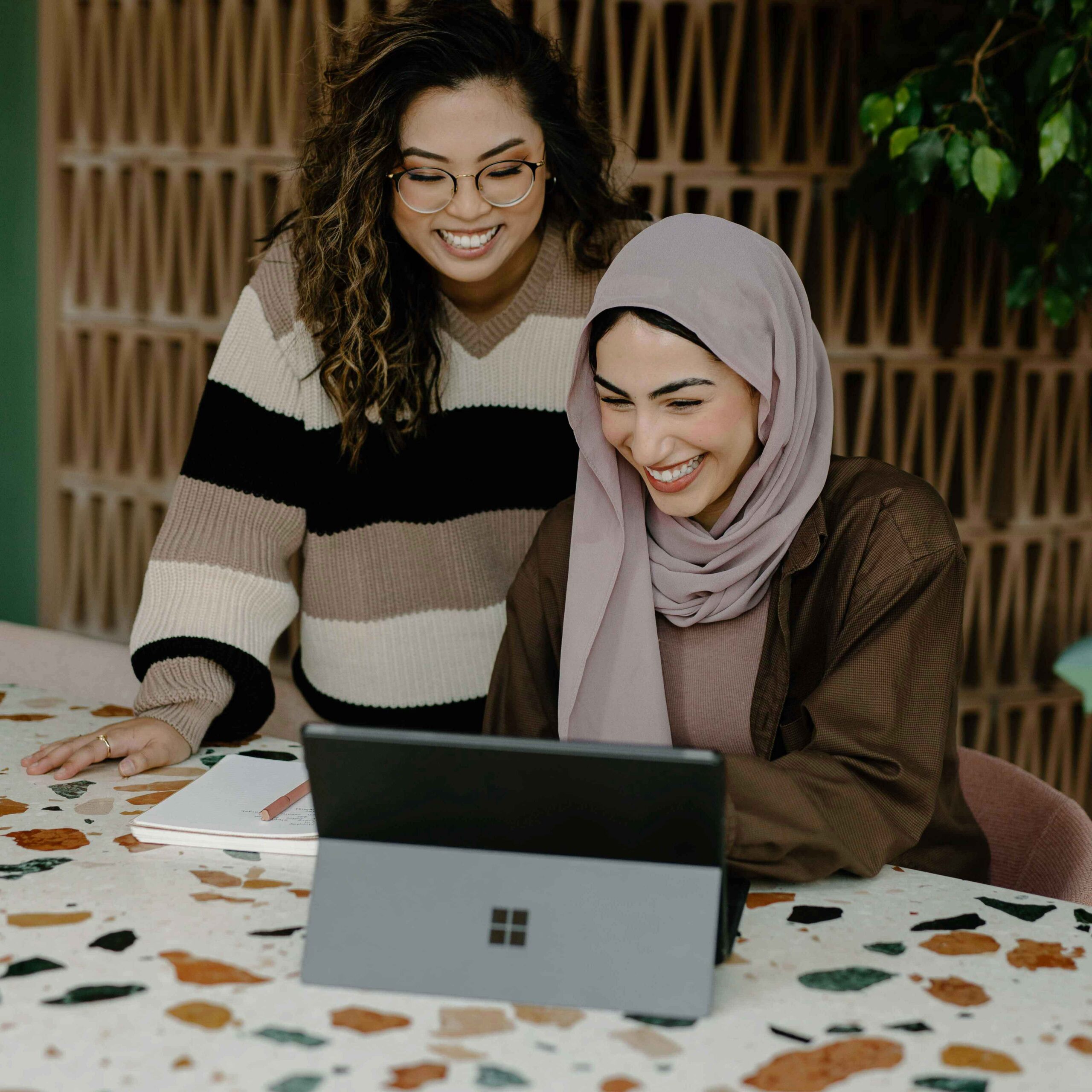 Two women smiling and working on a laptop at a terrazzo table. One wears glasses and a striped sweater, the other a hijab and brown shirt. Relaxed atmosphere.