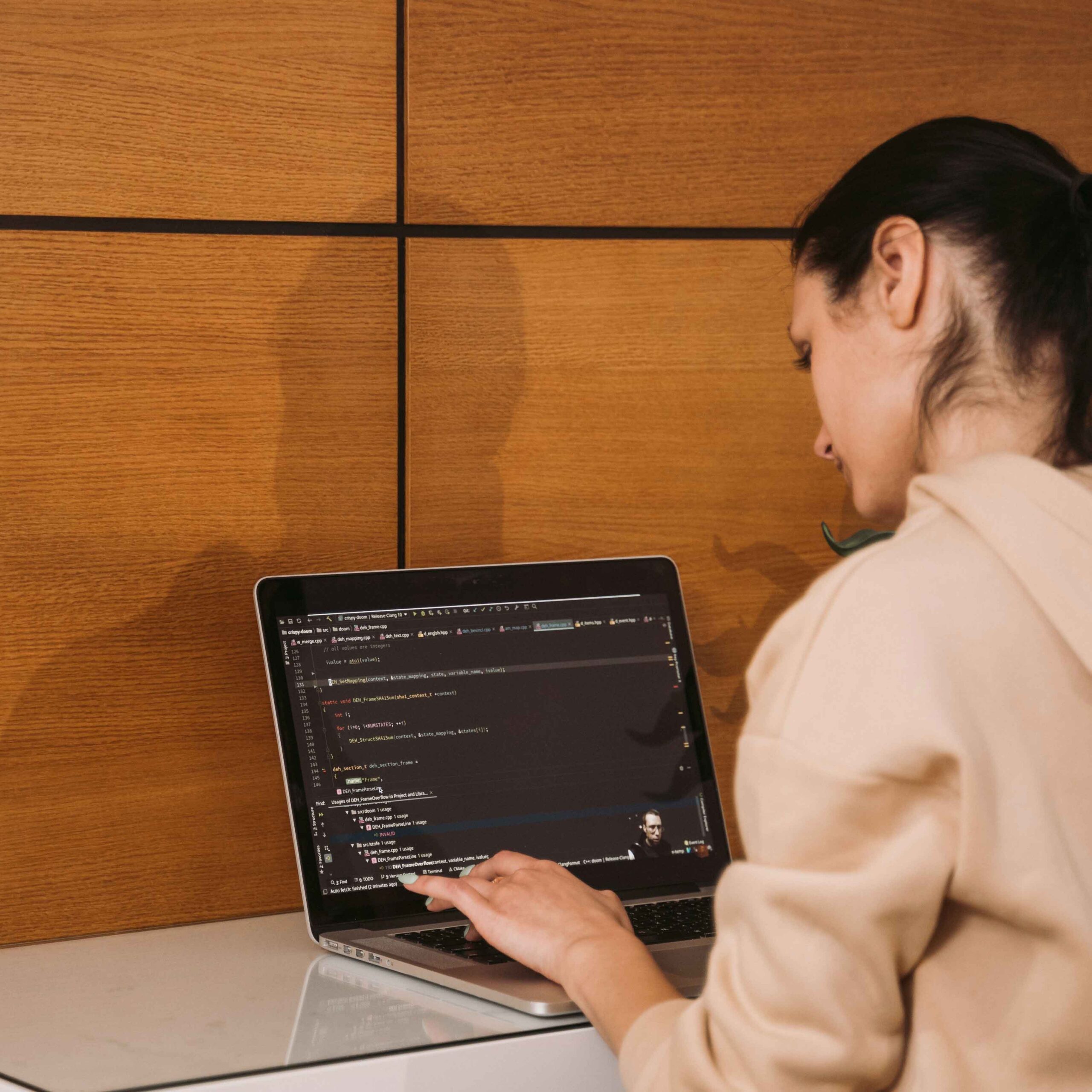 A person in a beige hoodie works on a programming code on a laptop against a wooden wall. The focus is on the coding, conveying concentration.