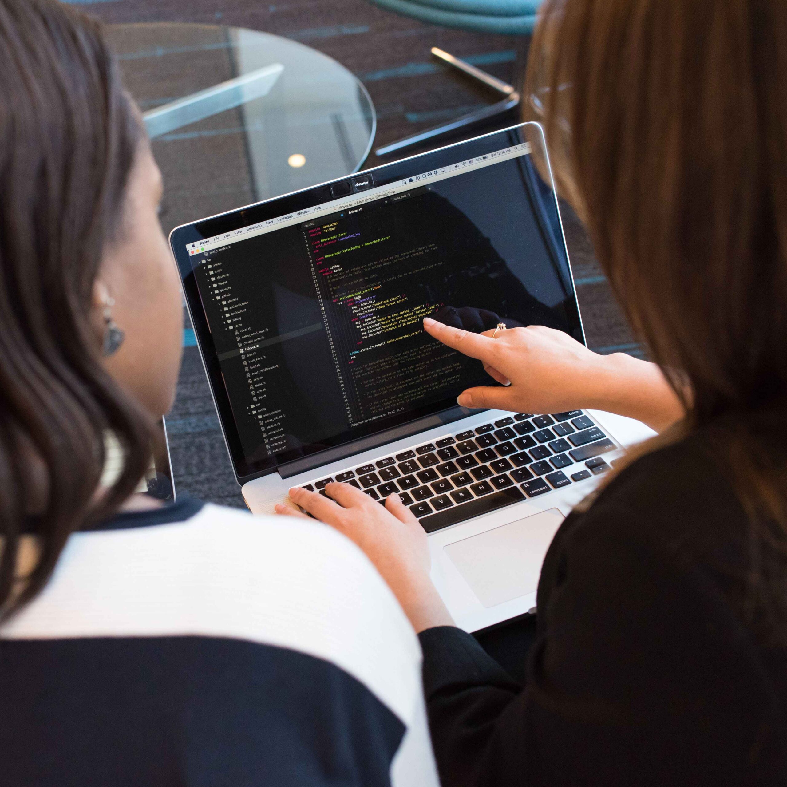 Two women work together, looking at a laptop screen displaying code. One points at the screen, suggesting collaboration and focus.