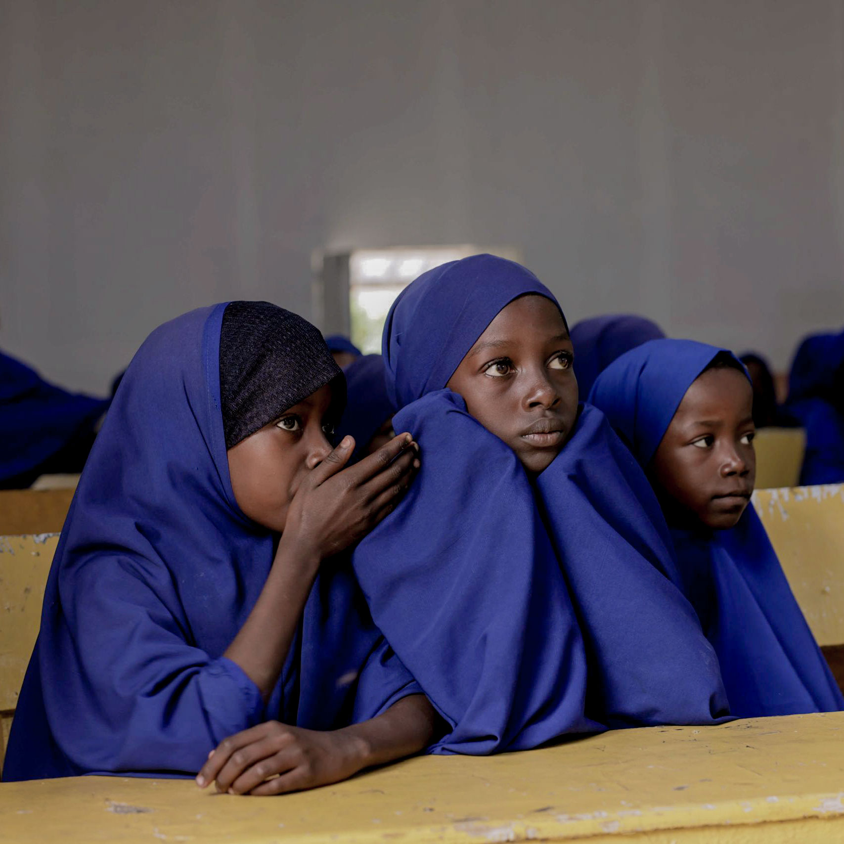 Three young girls in a classroom, wearing blue hijabs, sit at yellow desks. One whispers to another, creating a focused, contemplative atmosphere.