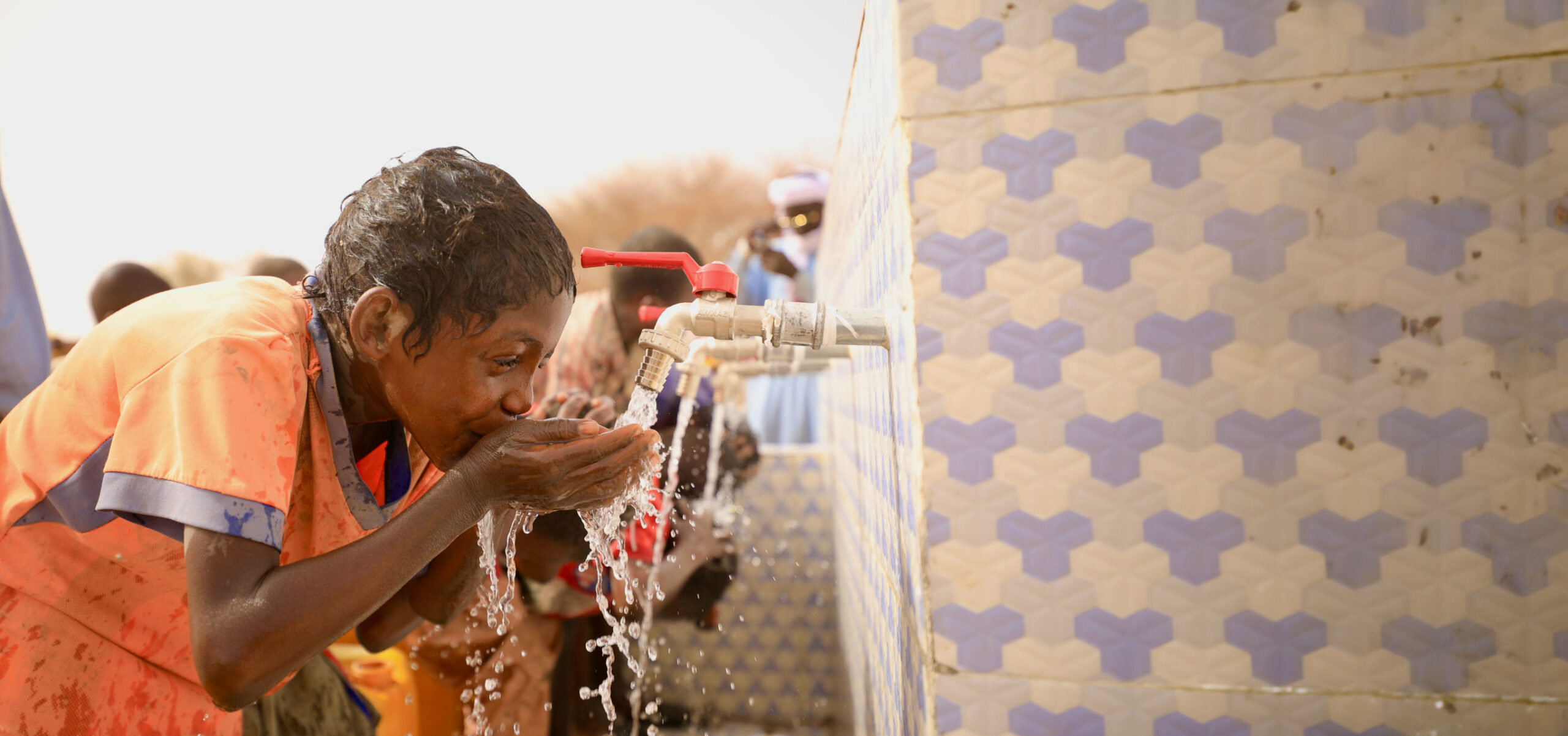 A boy in an orange shirt drinks water from a tiled fountain under a bright sky. The scene conveys relief and joy, with water splashing abundantly.