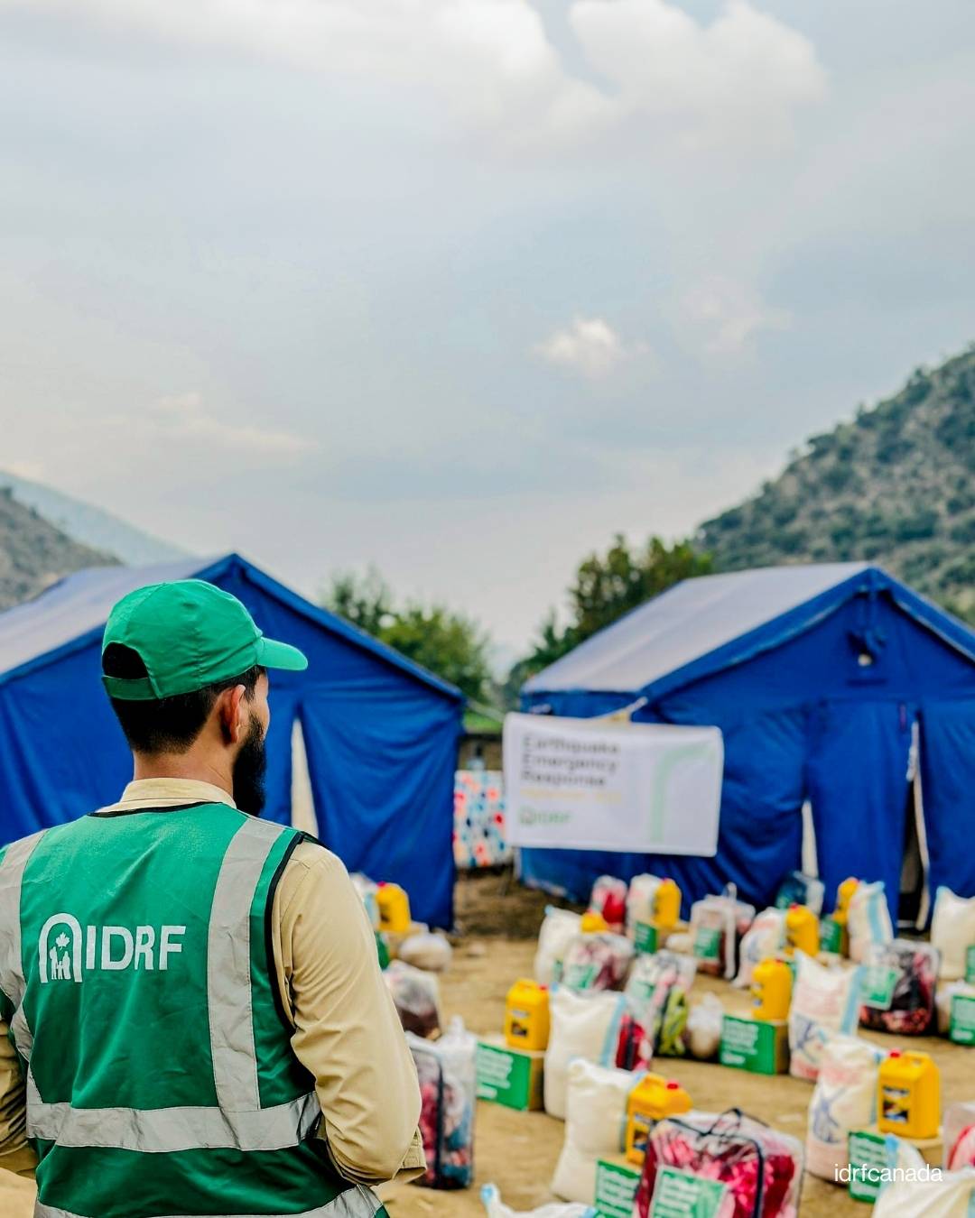 A man in a green vest stands beside a large pile of food, symbolizing generosity and community support.