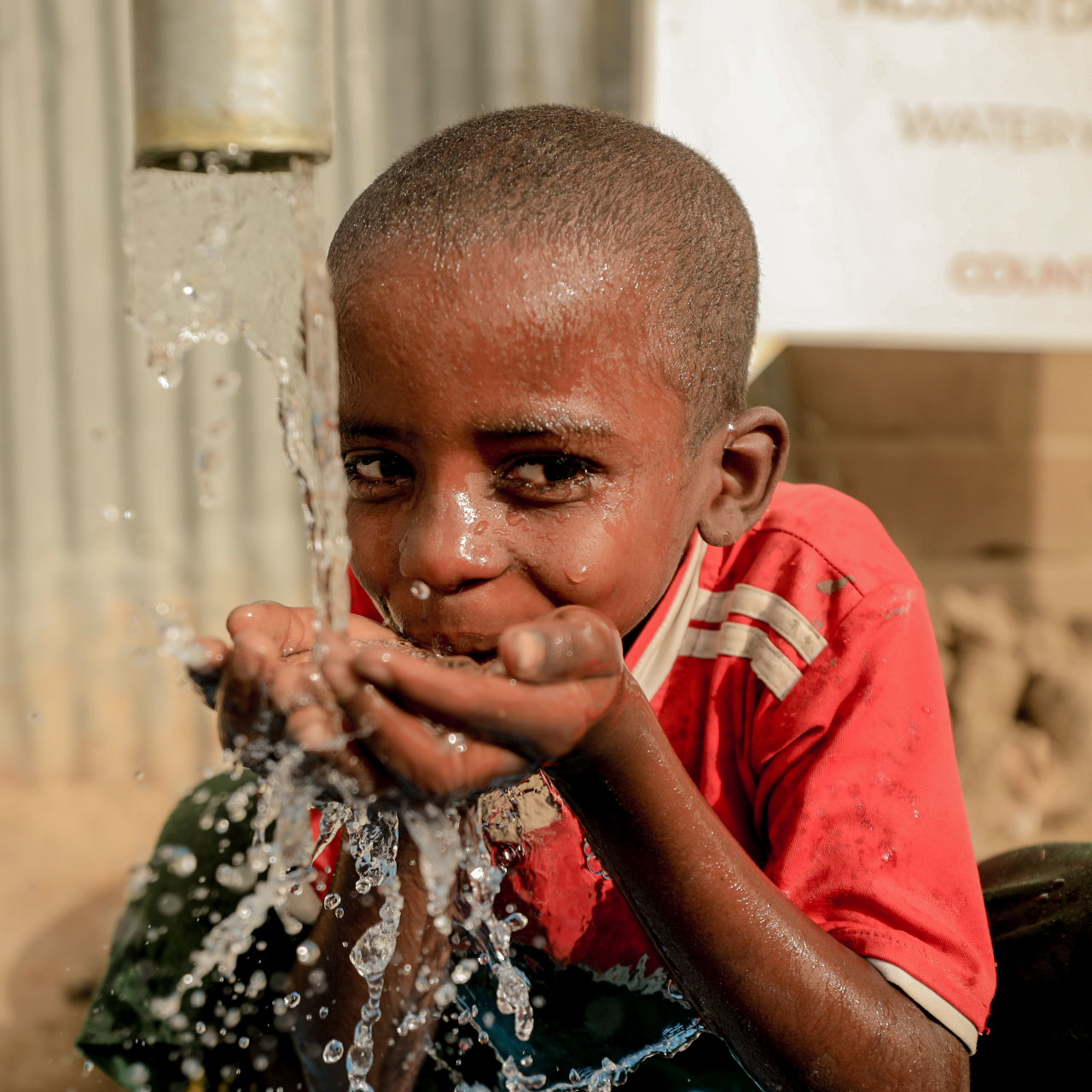 A young child in a red shirt gleefully drinks water from a pipe, cupping hands to catch splashing droplets. The scene conveys joy and refreshment.