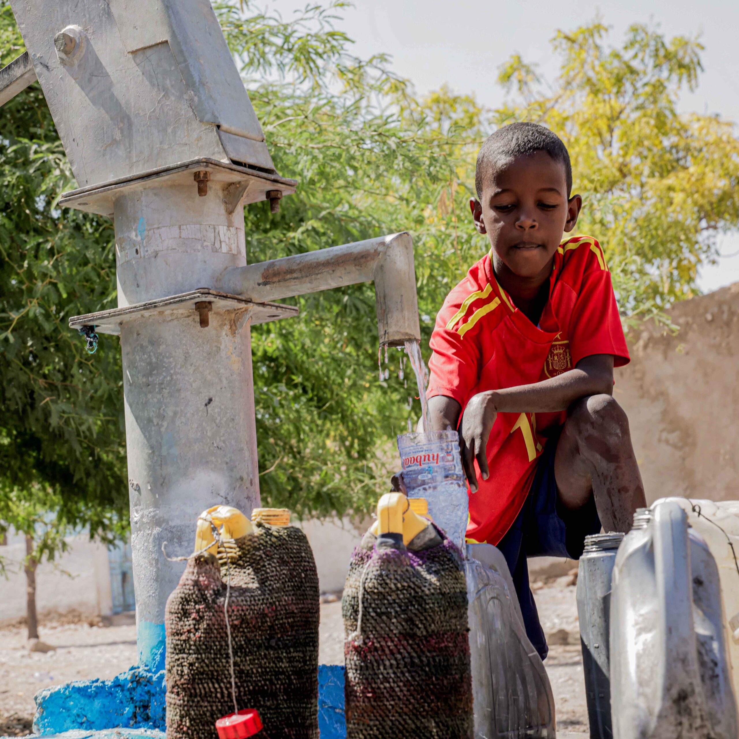 A boy in a red sports jersey fills a plastic bottle with water from a pump. Several jerry cans are nearby, with trees and a bright sky in the background.