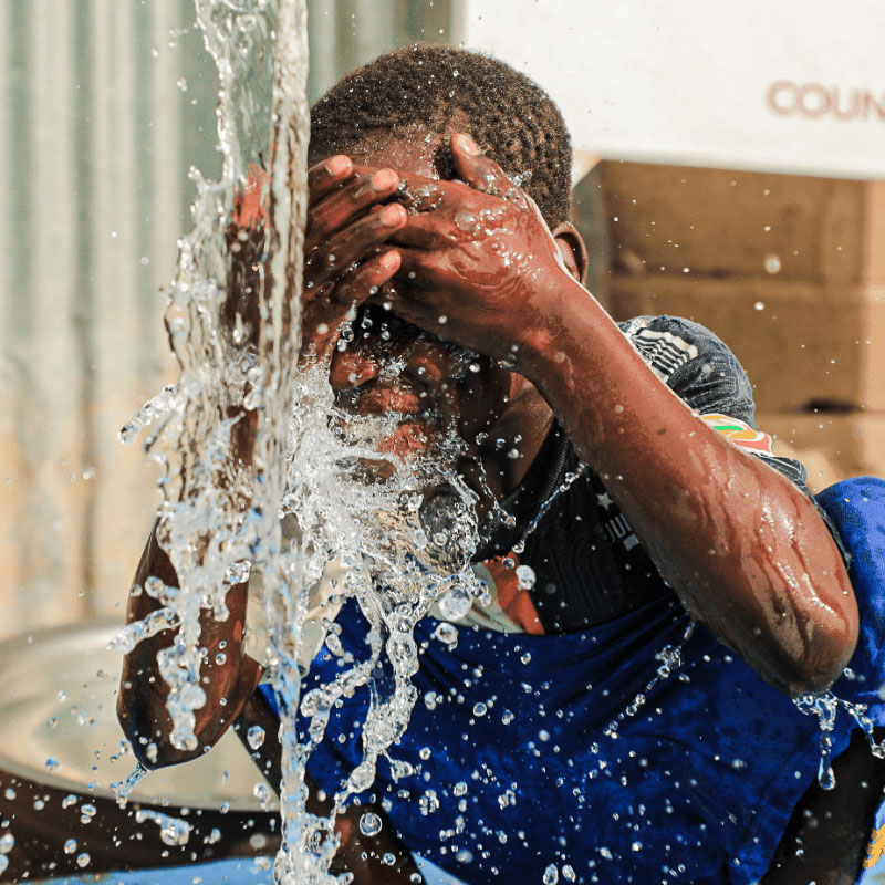 A person splashes water on their face from a flowing spout, with droplets suspended mid-air. The scene conveys refreshment and relief in a sunny setting.