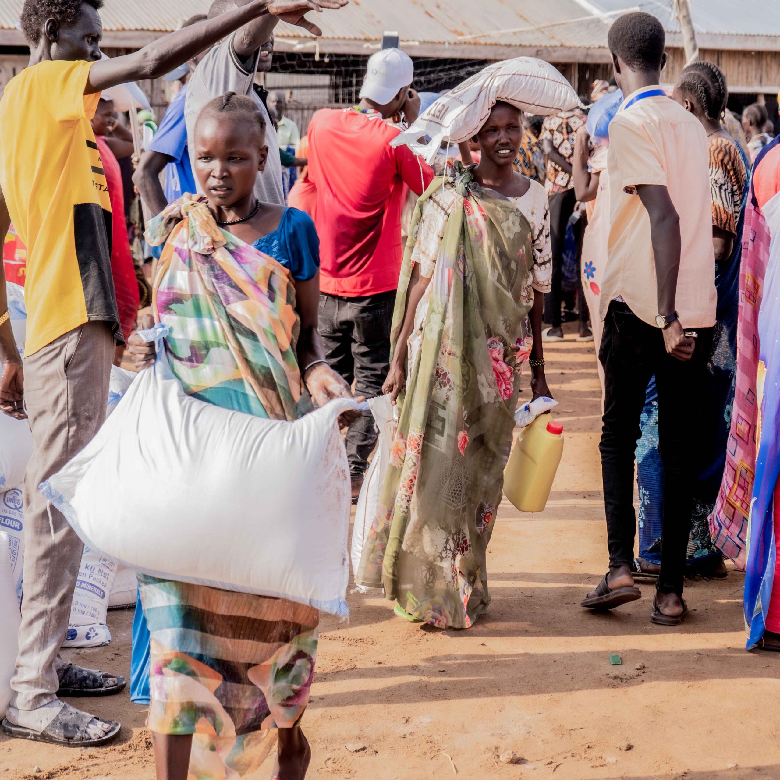 People in a crowded outdoor setting carry large bags on their shoulders. The atmosphere is busy, with colorful clothing and a sense of movement.