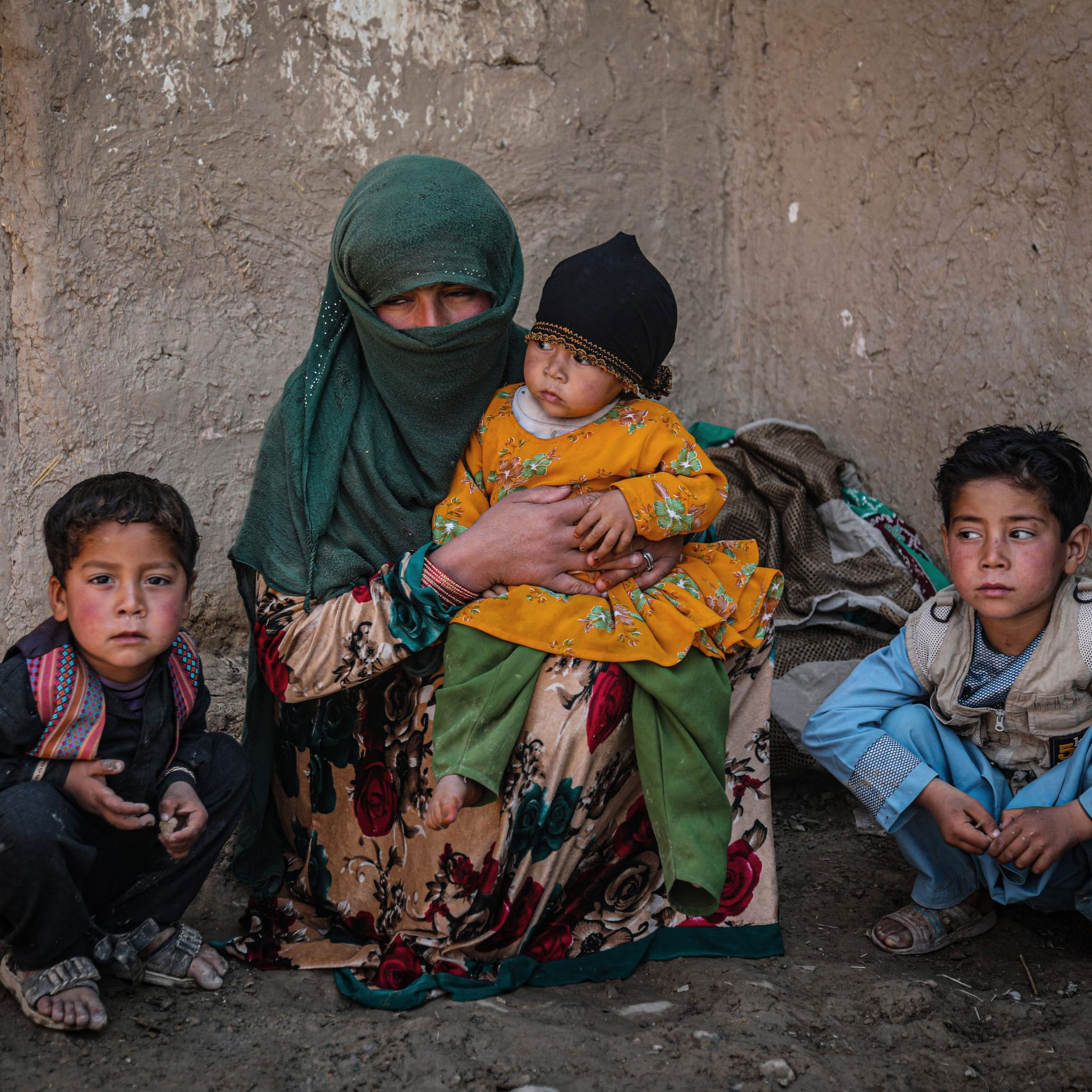 A woman in a green headscarf sits against a weathered wall, holding a child in yellow. Two boys sit with her, conveying a somber, resilient mood.