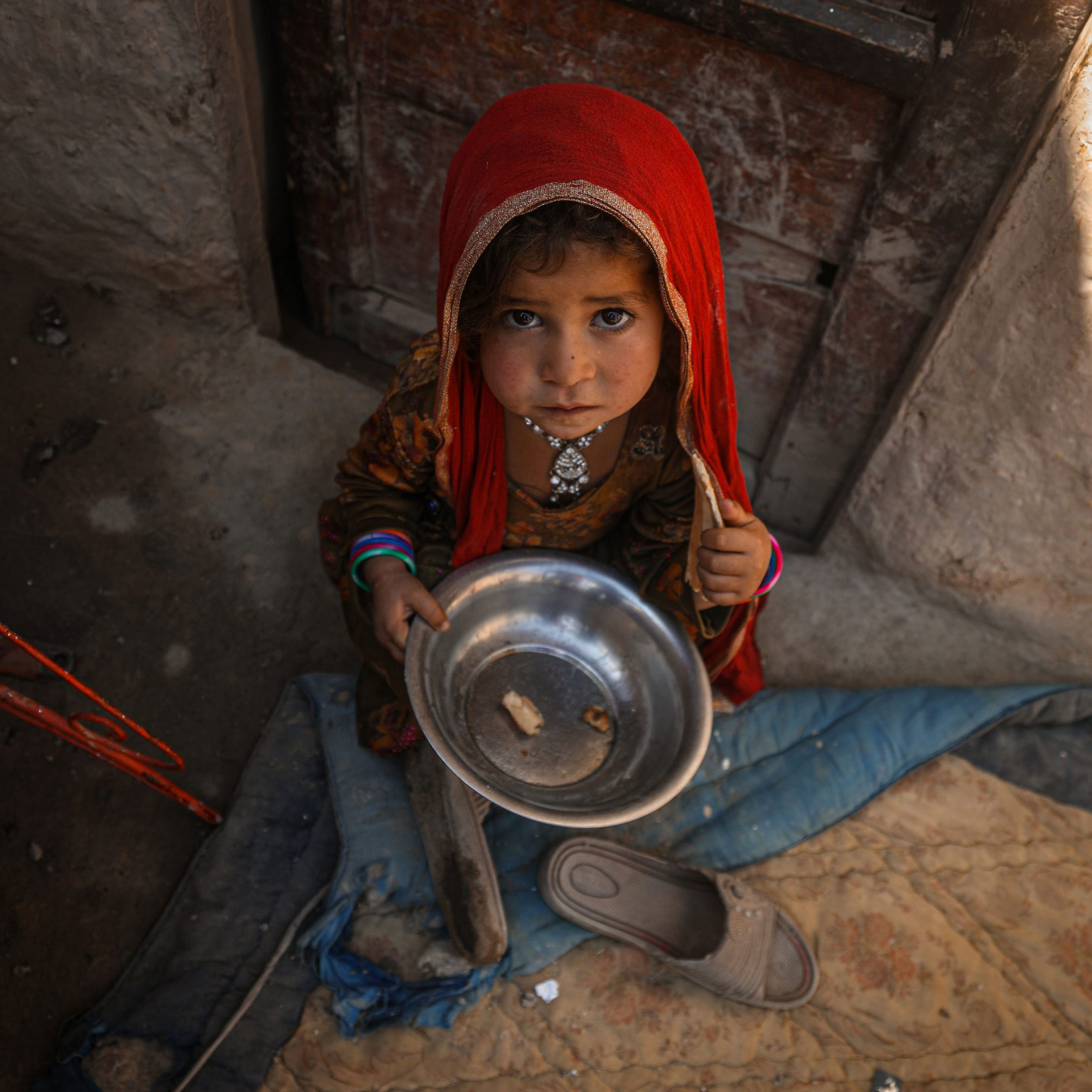 A young child with a solemn expression, wearing a red headscarf and colorful bracelets, holds an empty metal bowl while sitting on a mat near a shoe.
