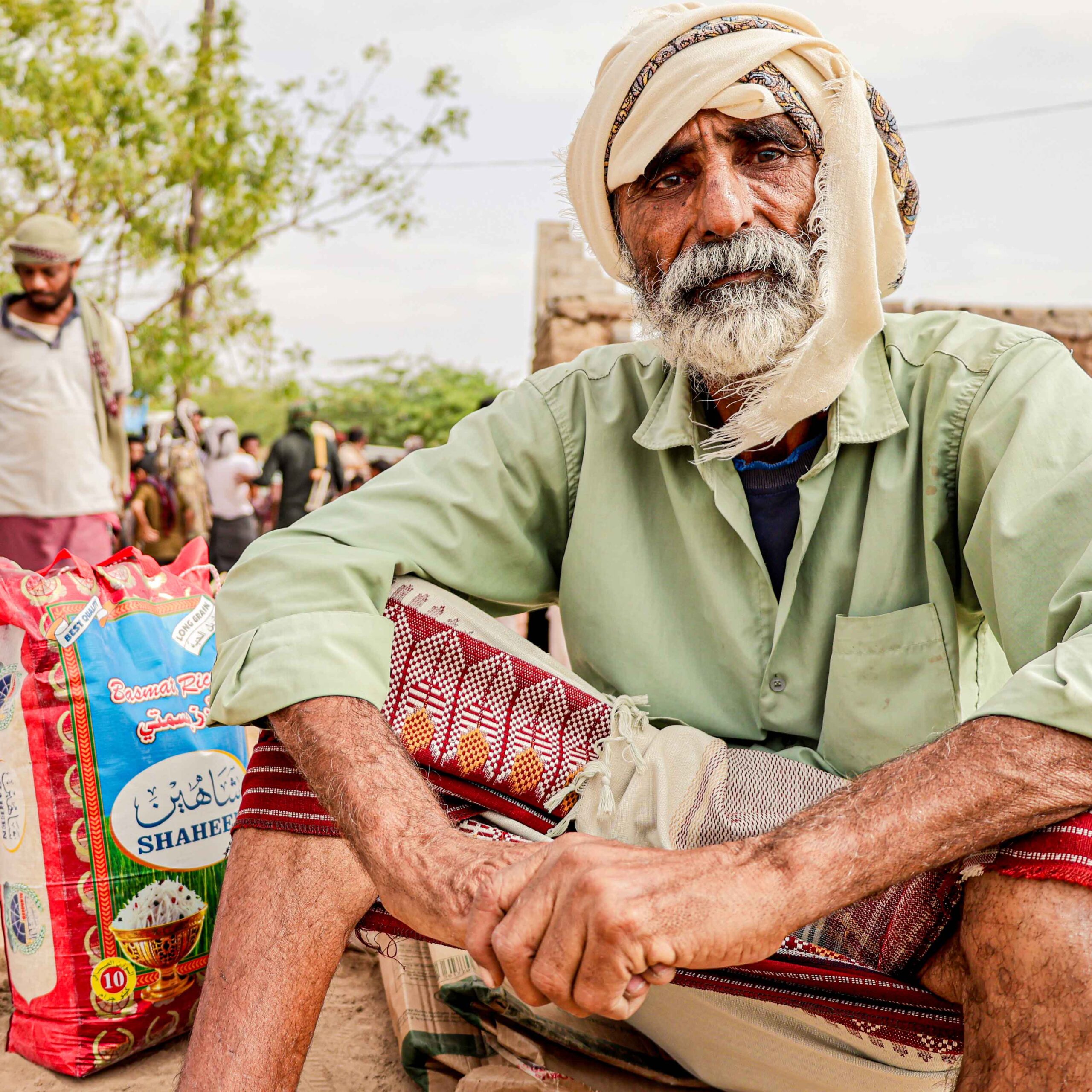 An elderly man with a weathered face and a neutral expression sits outdoors beside a colorful bag labeled "Shaheer." He wears a light green shirt and a beige turban.