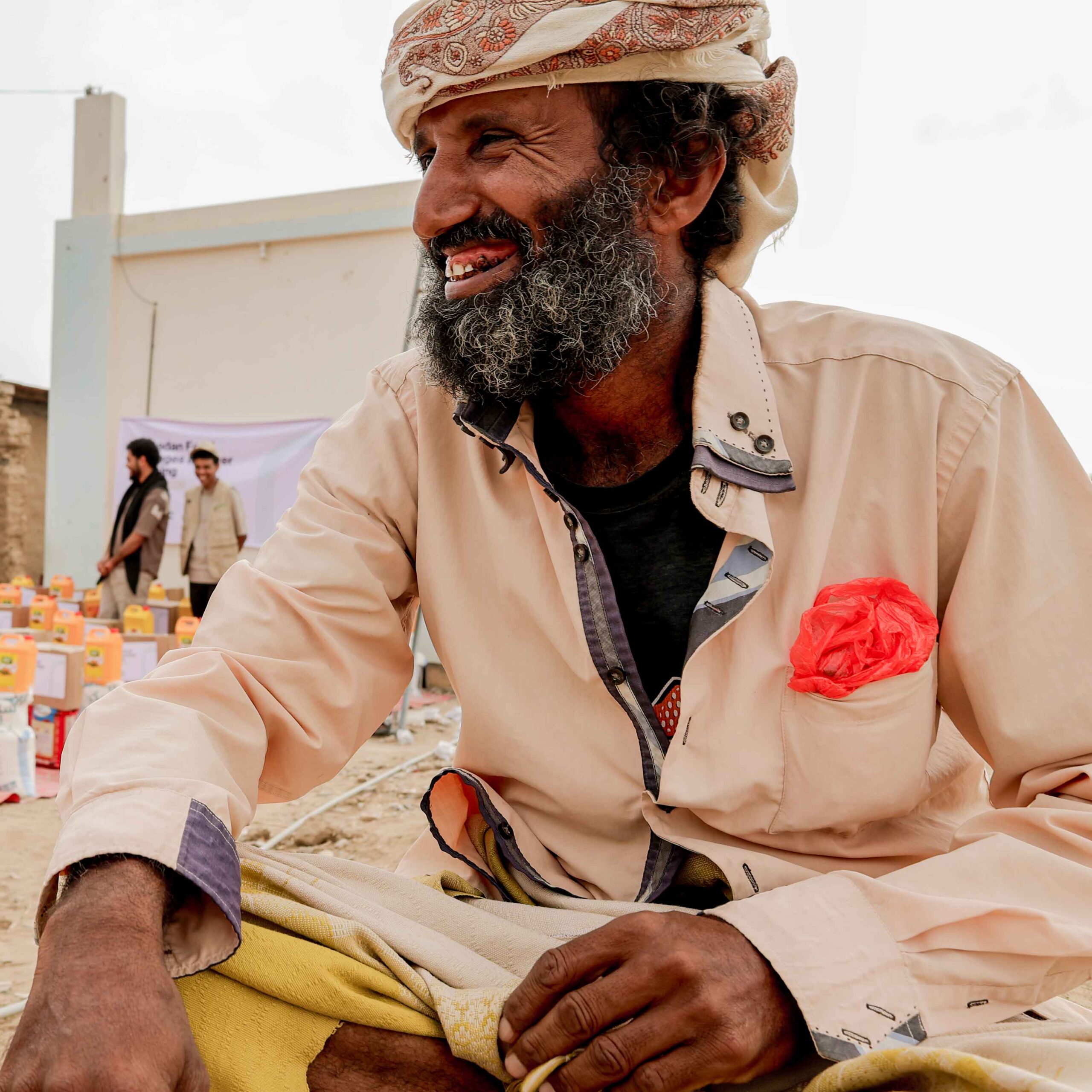 A bearded man smiles, wearing a headscarf and pale shirt with a red pocket handkerchief. In the background, aid supplies and people are visible.