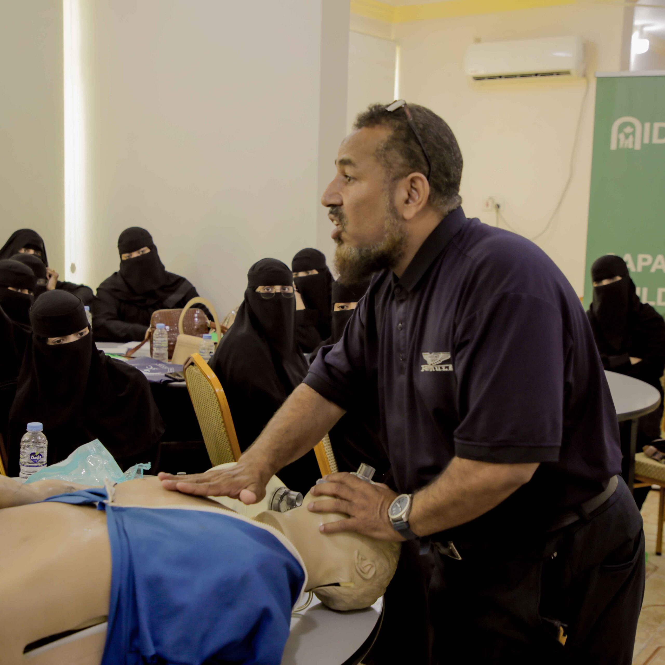 Instructor demonstrating CPR on a mannequin to a group of women wearing niqabs in a classroom setting. The mood is focused and educational.
