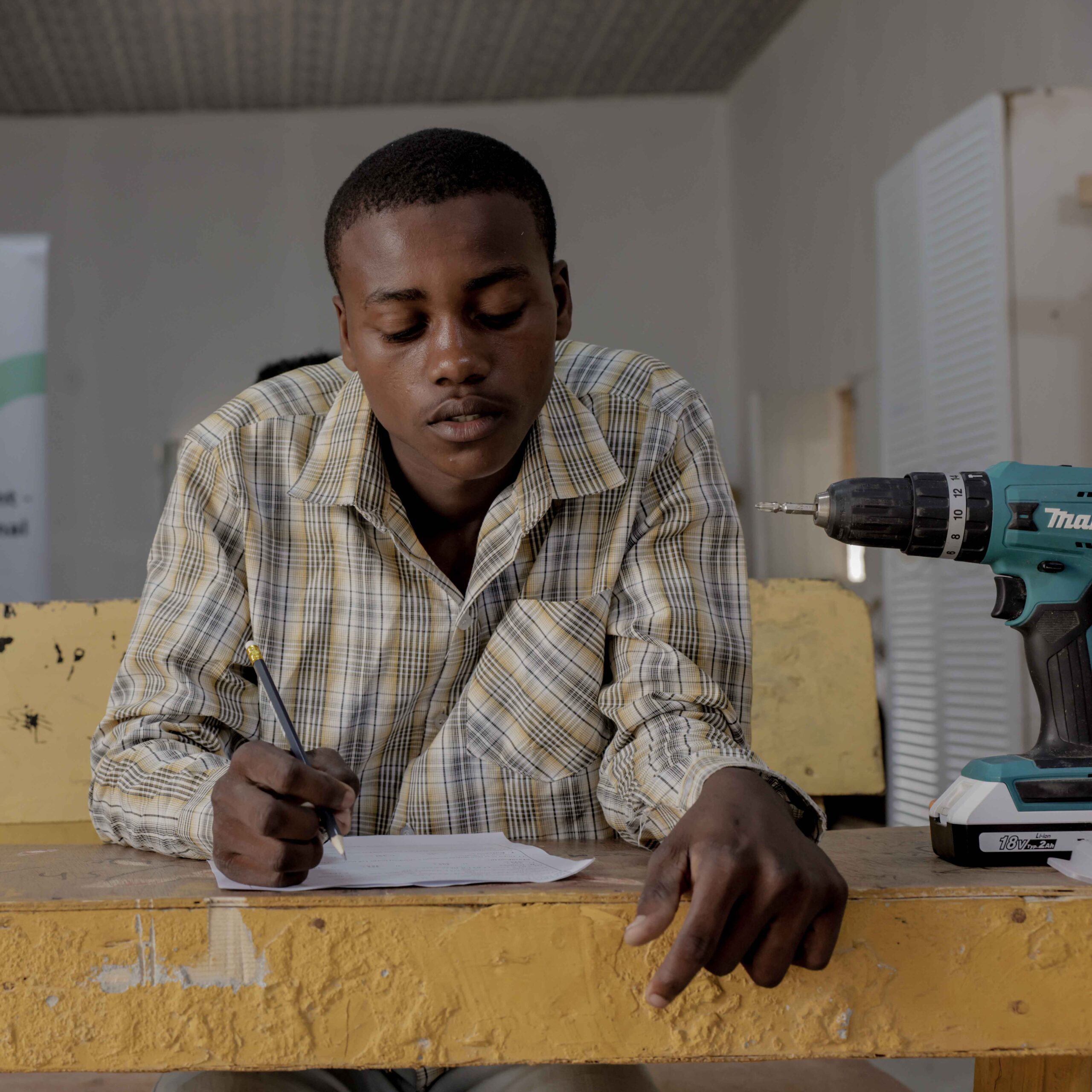 A man sits at a table, focused on a drill and a piece of paper, possibly working on a project or repair.