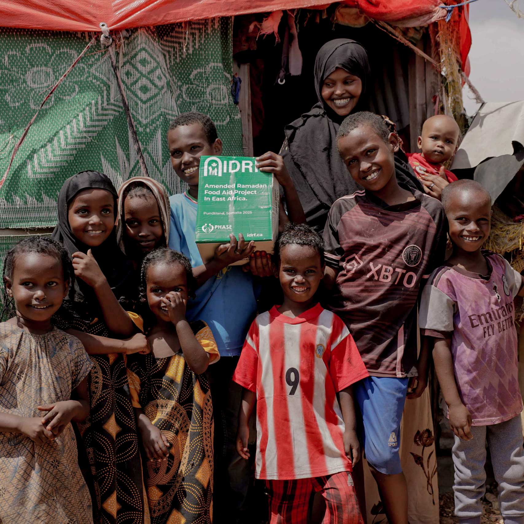 A group of smiling children stands together outdoors, holding a sign about a Ramadan food aid project in Somalia. A sense of joy and community is conveyed.