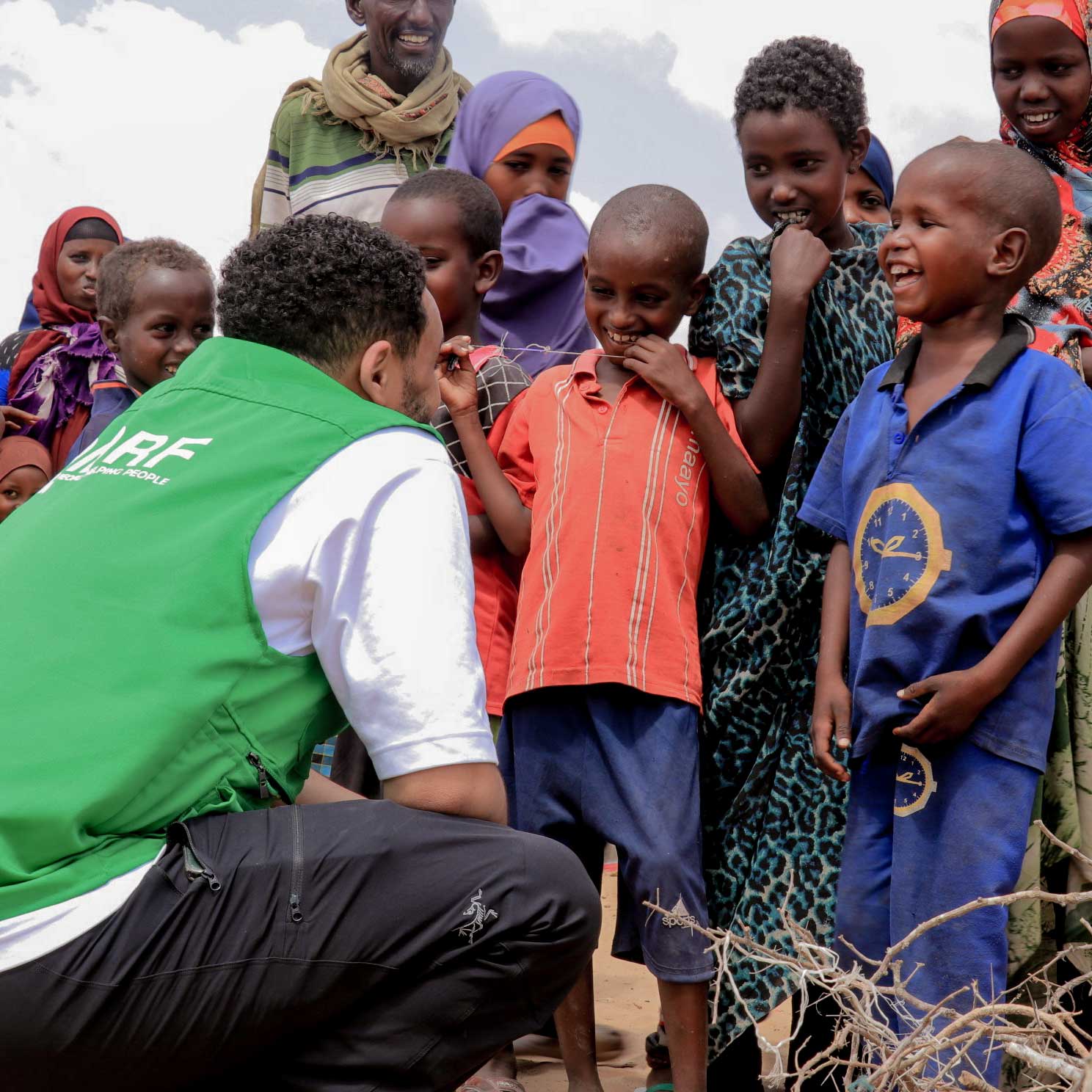 A person in a green vest engages with smiling children in colorful clothing, conveying a sense of joy and community under a bright sky.