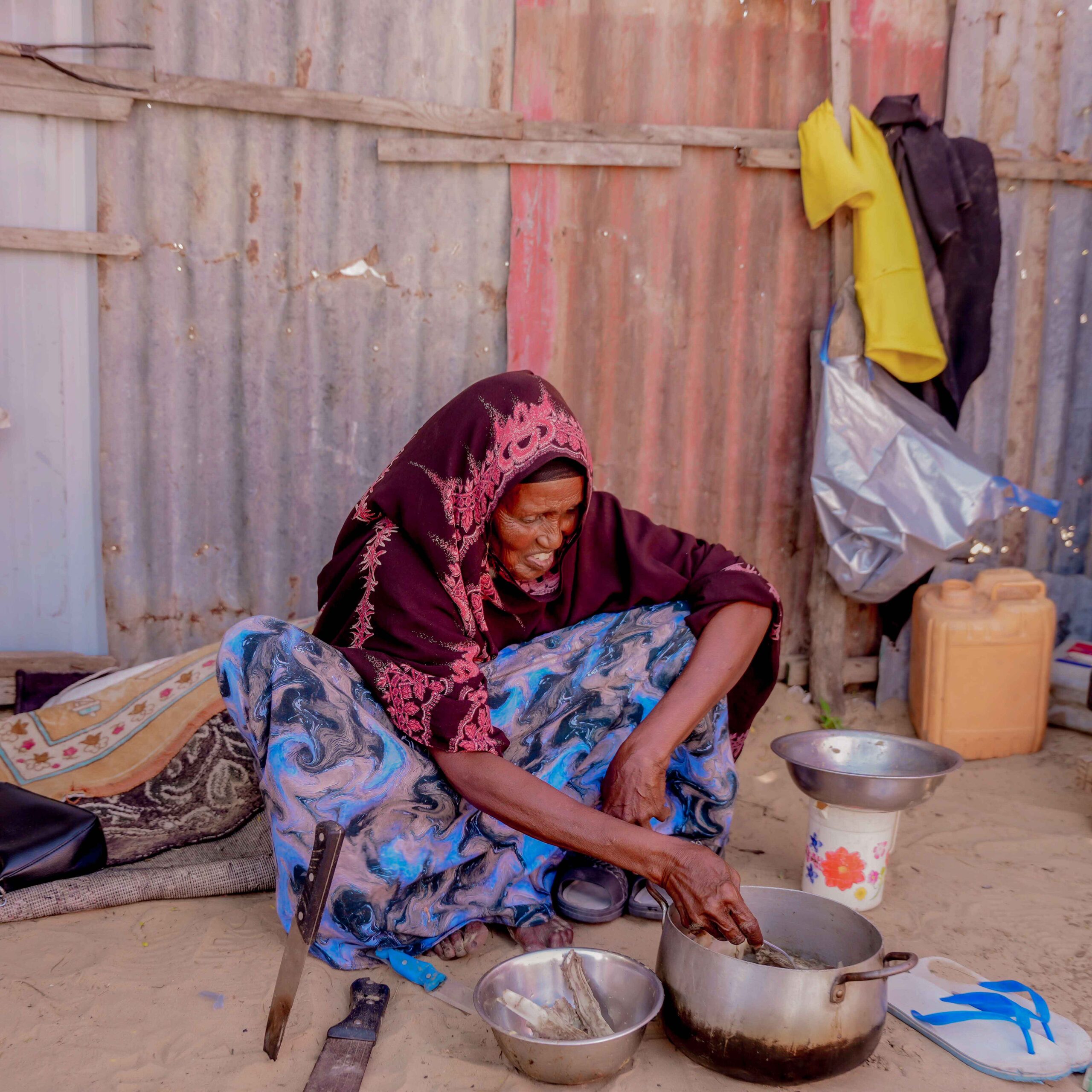 A woman cooking, surrounded by sand, with ingredients laid out around her.
