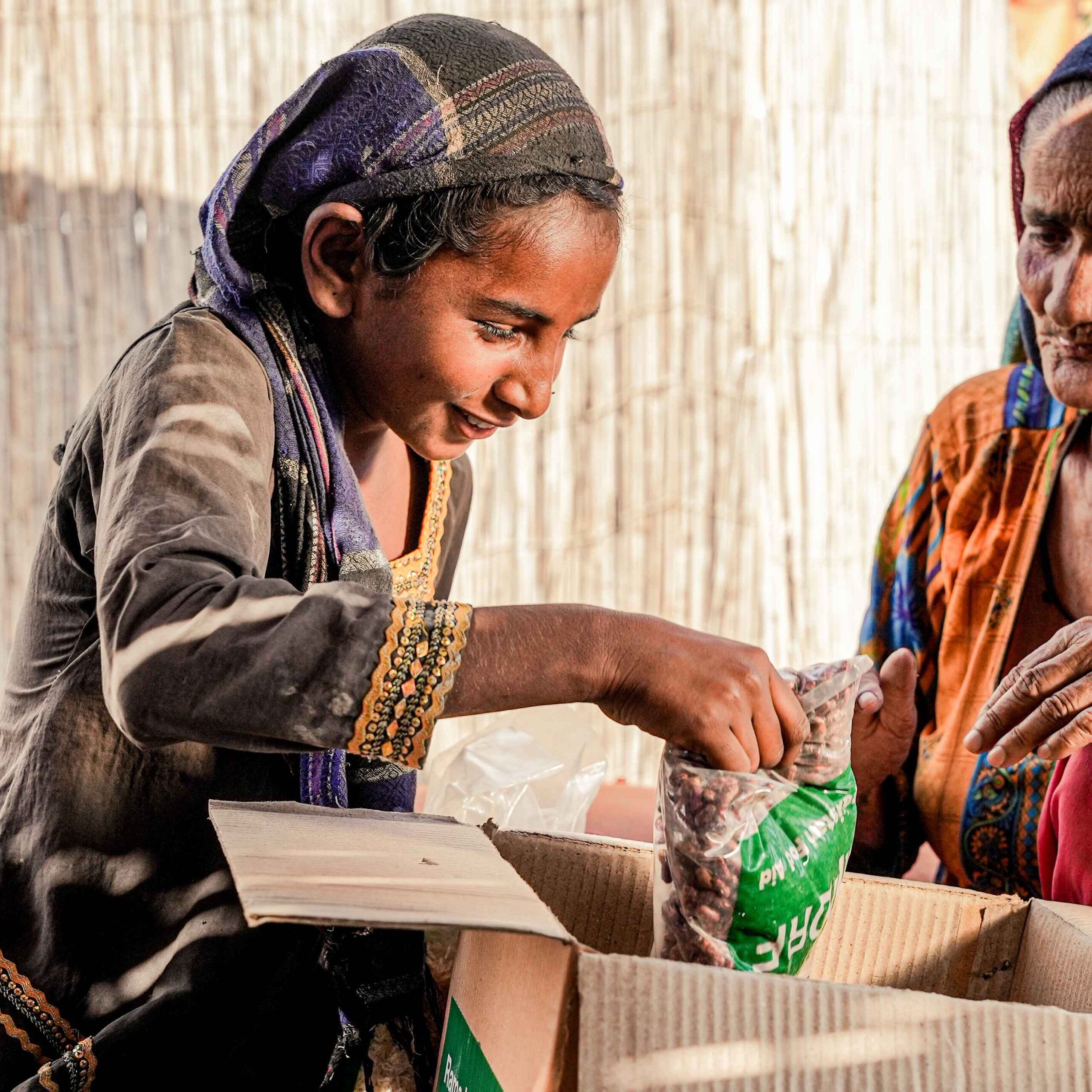 A smiling child in traditional attire eagerly examines a bag of food from an open box, where an older woman in colorful clothing looks on warmly.