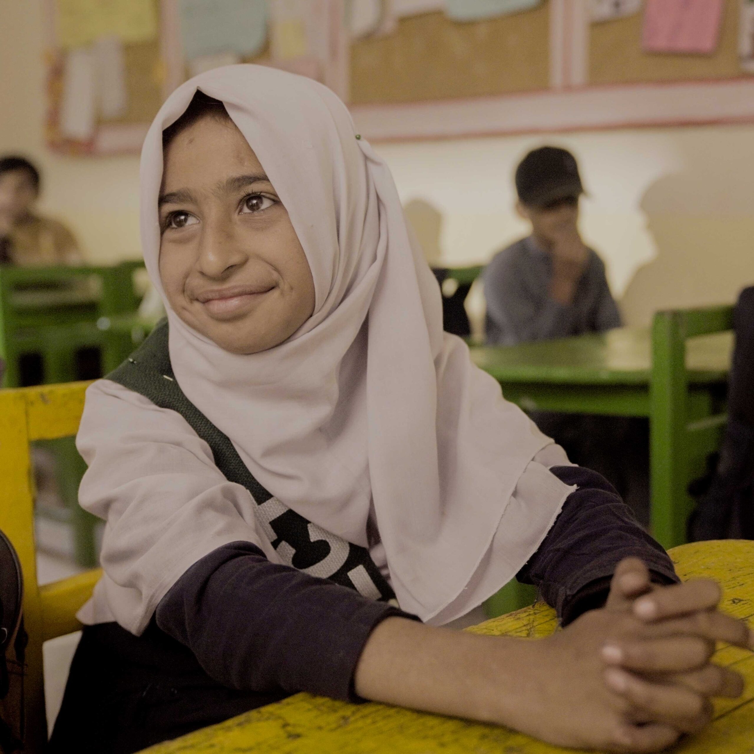 A young girl in a classroom wears a light hijab, smiling softly. She sits at a yellow desk with a thoughtful expression. Background shows students and a bulletin board.