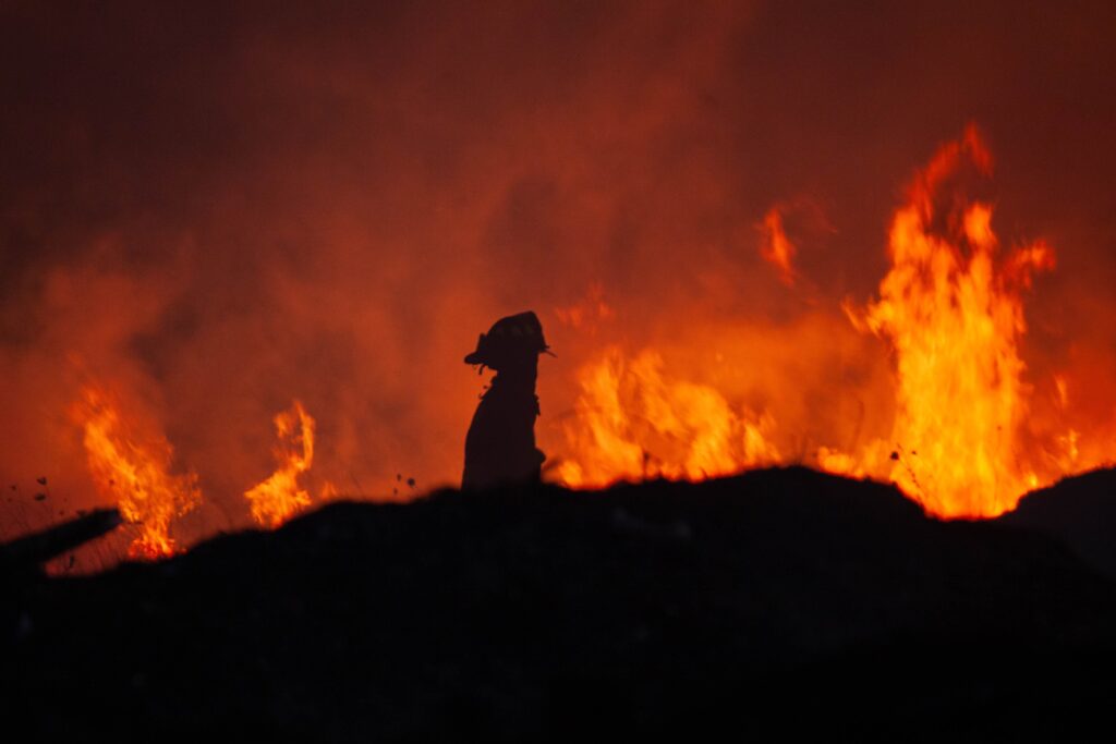 A firefighter stands confidently in the center of a large fire, surrounded by flames and smoke.