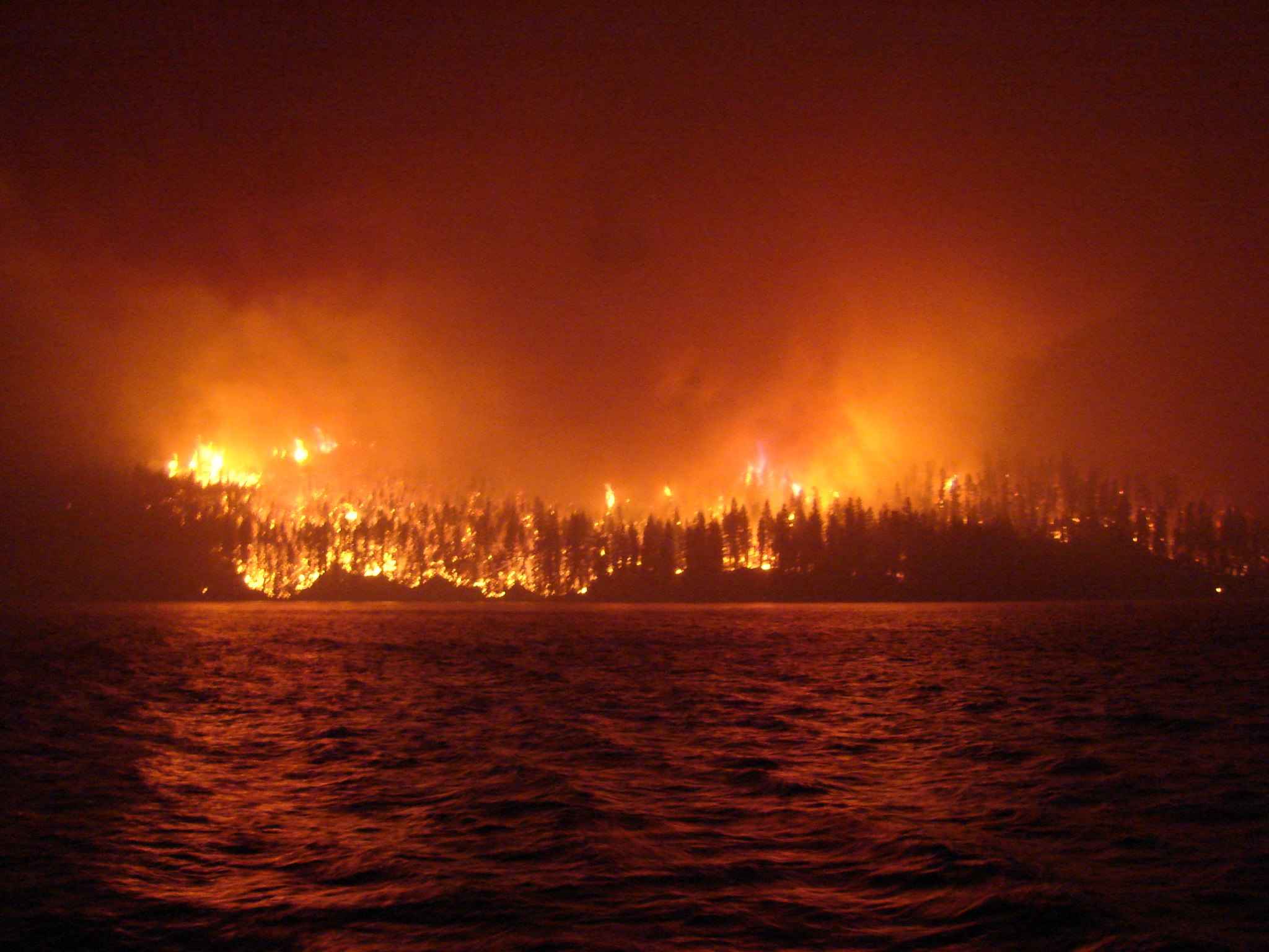 A fire burns brightly on the shore of a tranquil lake, with flames reflecting on the water's surface.